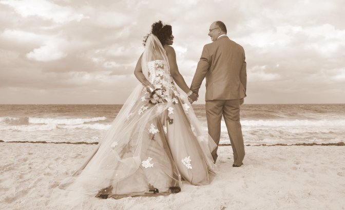 Newlyweds, a Black bride in a strapless wedding dress with floral embellishments and a groom in a suit, hold hands on a sandy beach, gazing out at the ocean under an overcast sky in a sepia-toned photo.