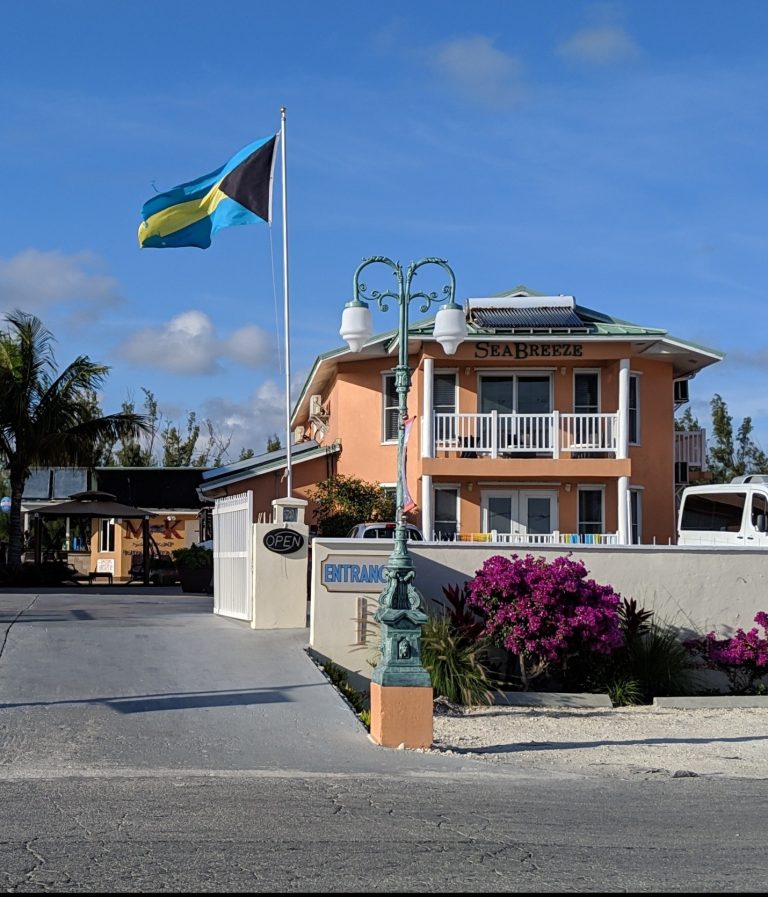 A peach-colored building labeled "SEABREEZE" with a balcony, surrounded by a white fence and vibrant pink flowers. A Bahamas flag flies on a pole, and a vintage-style lamp post is nearby. An "OPEN" sign and "ENTRANCE" sign are visible by a driveway under a clear blue sky.